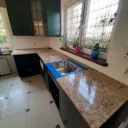 Kuchnia z ciemnozielonymi szafkami i blatem z brązowego kamienia, z widocznym zlewem zabezpieczonym niebieską folią, oraz oknem z kwiatami i firankami w tle.