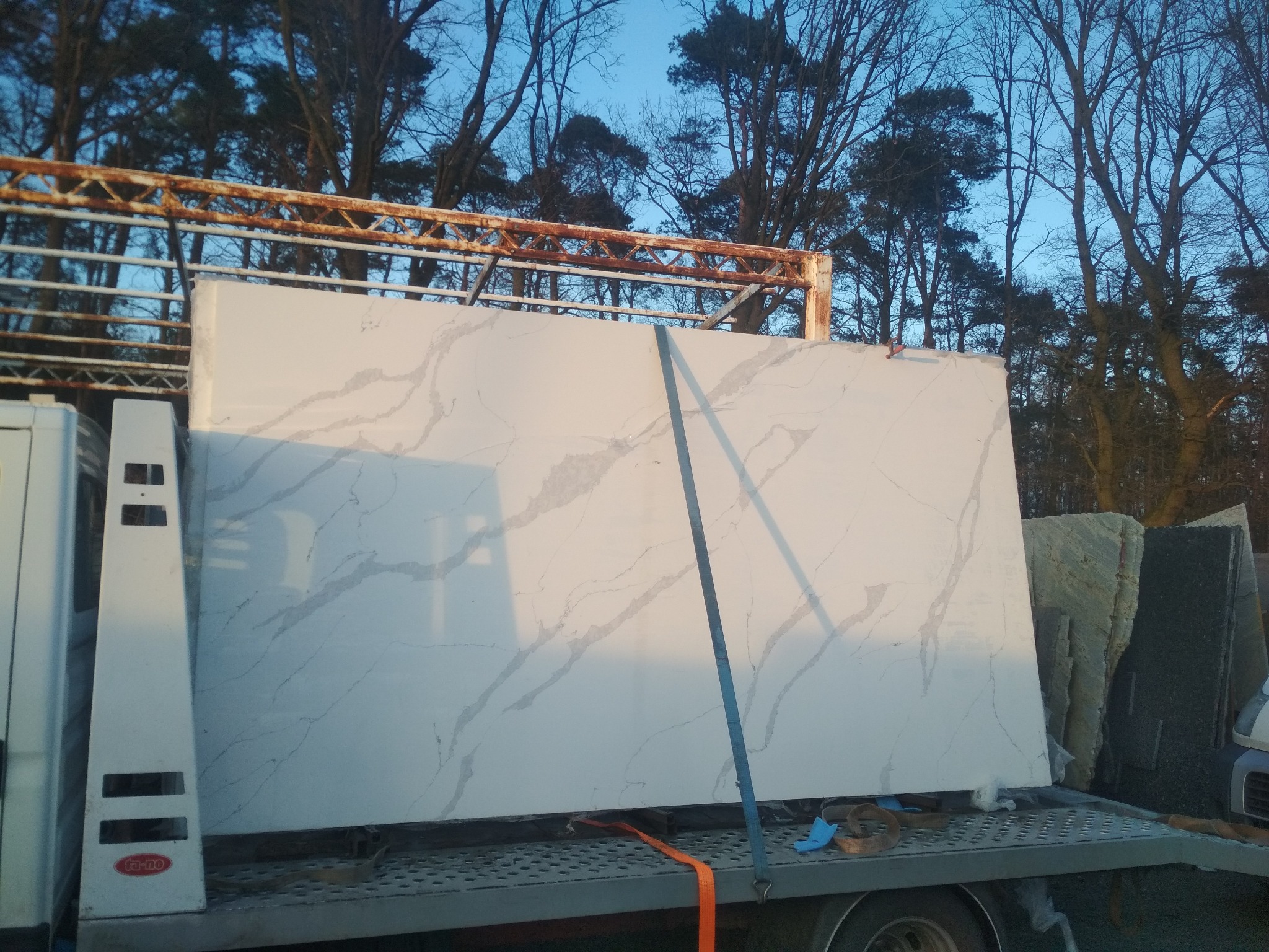 Duża płyta z białego kamienia z szarymi żyłkami, zabezpieczona pasami na przyczepie samochodu dostawczego, widoczne inne płyty kamienne i drzewa w tle.