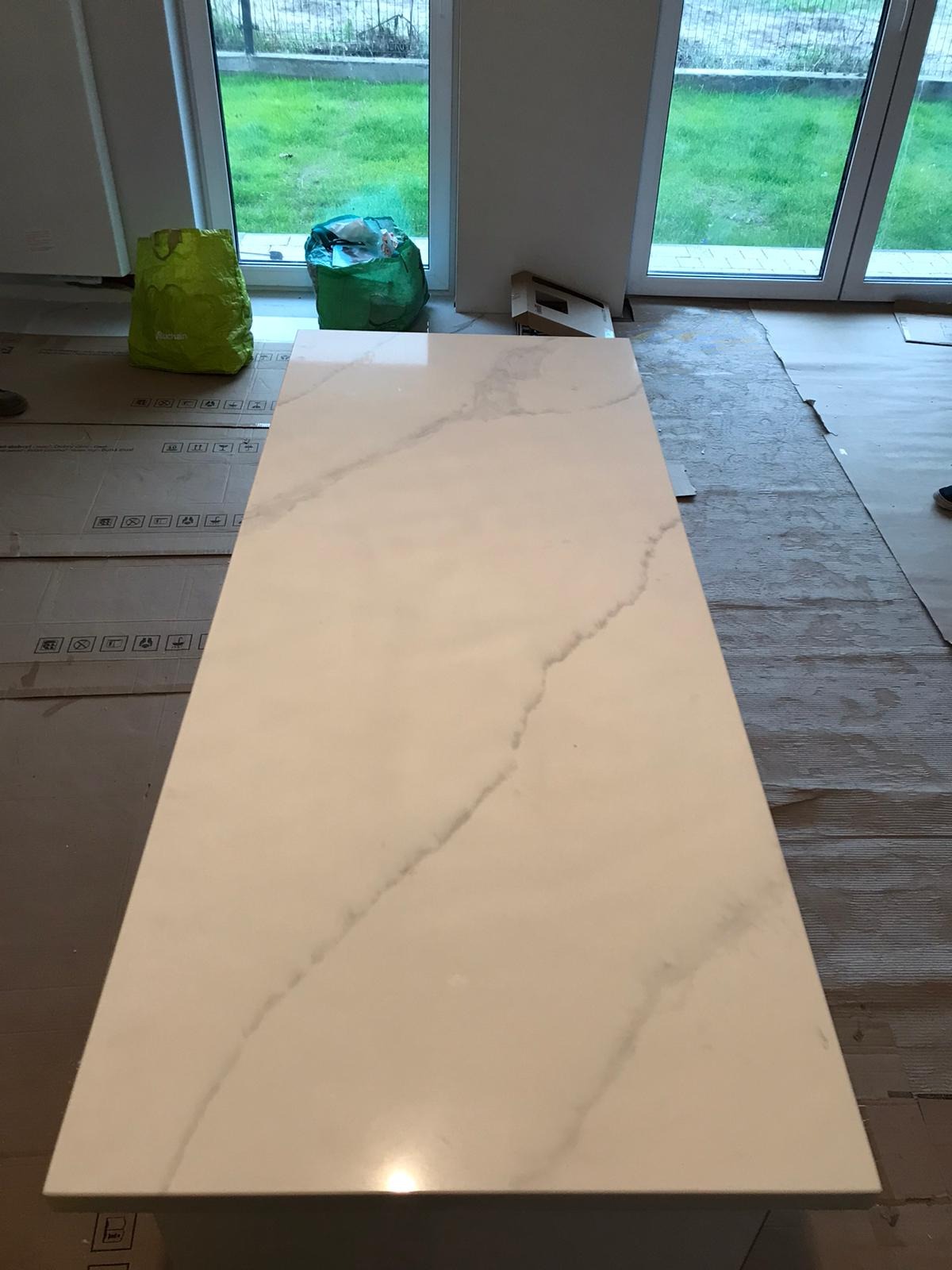 Biały blat z marmurowym wzorem, widoczne zabezpieczenia podłogi i okno z widokiem na trawnik w Strzegomiu, sugerujące prace wykończeniowe.