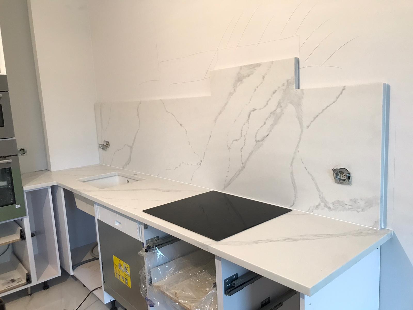 Montaż blatu kuchennego z białego marmuru z czarną płytą indukcyjną, widoczne niedokończone szafki i ślady ołówka na ścianie.