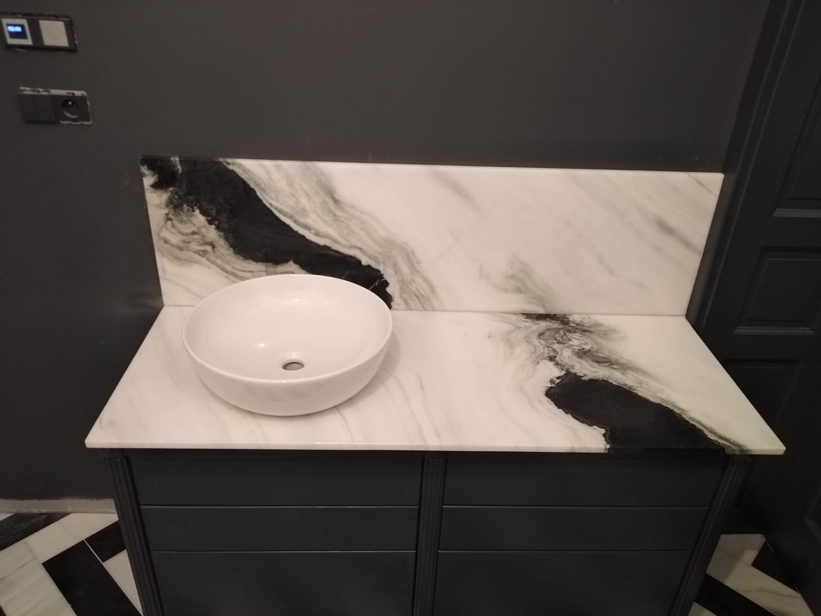 Biała umywalka nablatowa na blacie z płyty imitującej marmur z czarnymi żyłami, zamontowana na ciemnoszarej szafce łazienkowej.
