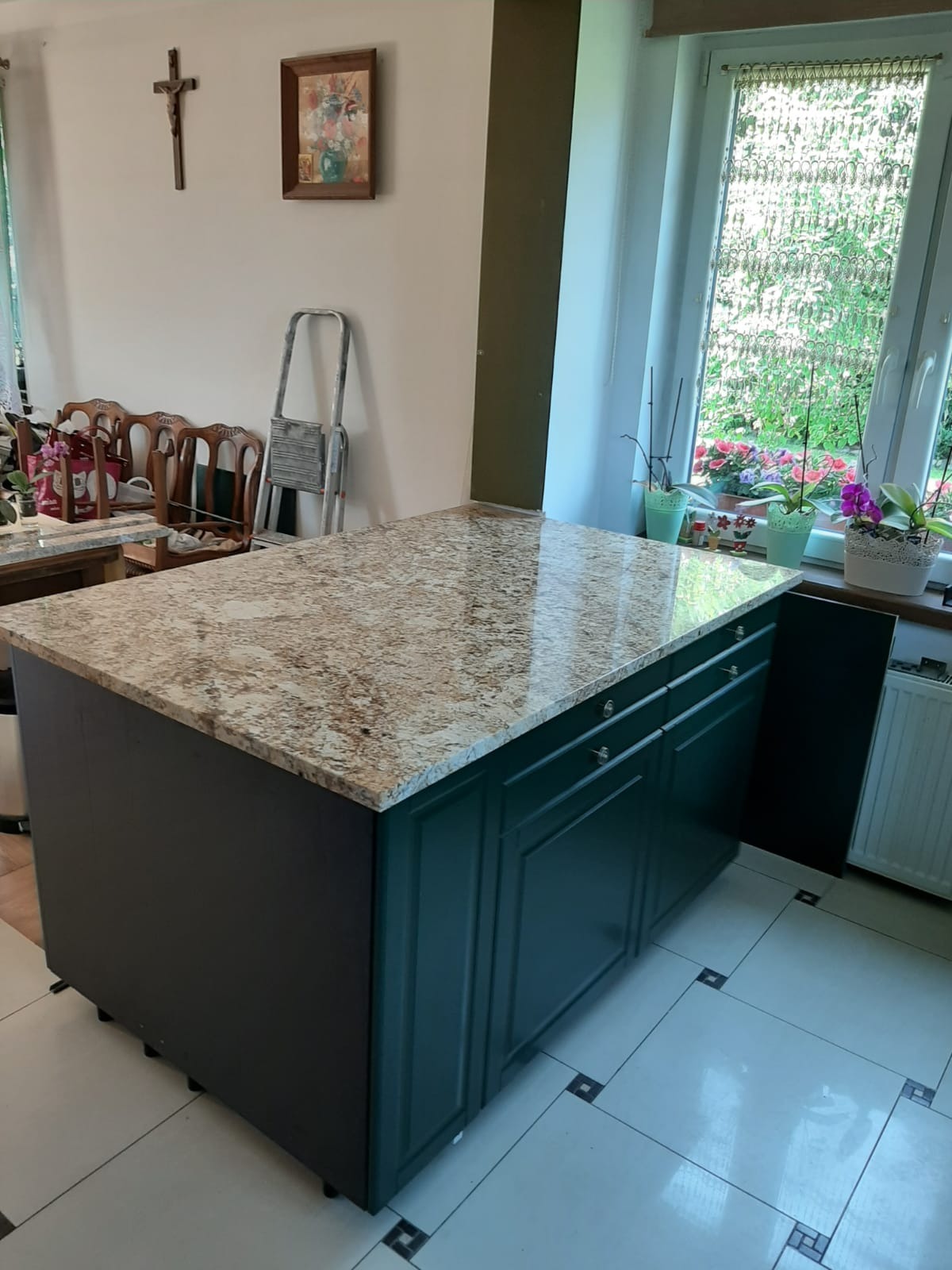 Wyspa kuchenna z blatem z kamienia o beżowo-brązowej teksturze, ciemnozielone szafki, widok na okno z kwiatami i krzyż na ścianie w tle.