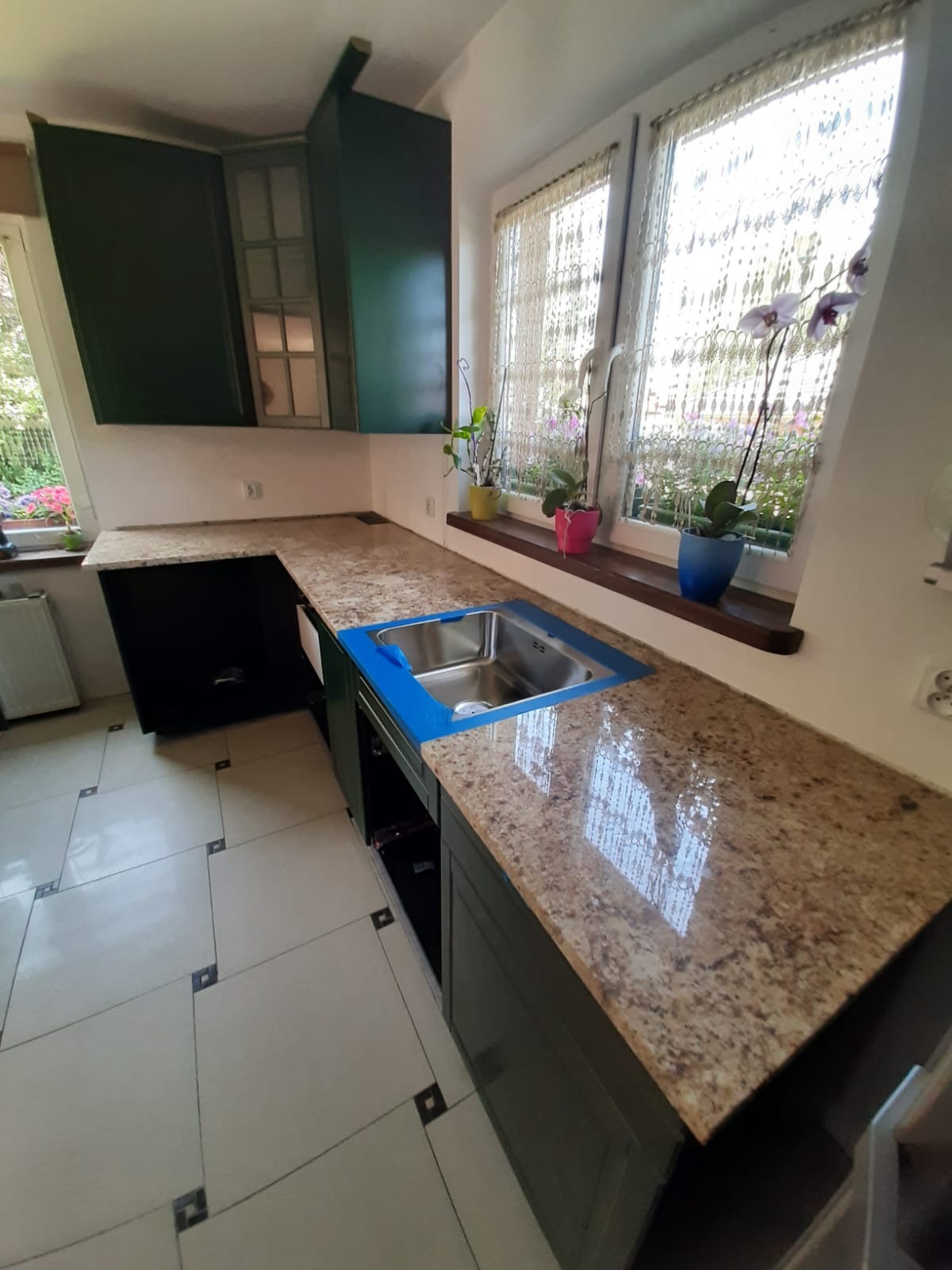 Kuchnia z ciemnozielonymi szafkami i blatem z brązowego kamienia, z widocznym zlewem zabezpieczonym niebieską folią, oraz oknem z kwiatami i firankami w tle.