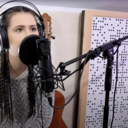 Wokalistka z warkoczami śpiewa do mikrofonu w studiu, w tle gitary i panel akustyczny z otworami.