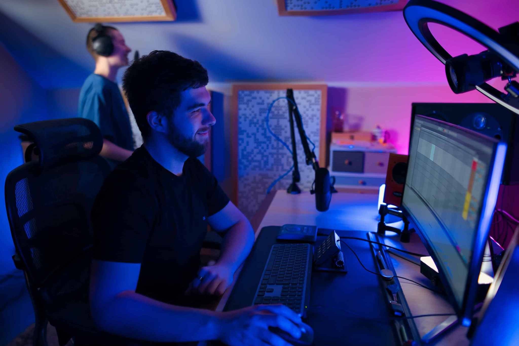 Młody mężczyzna uśmiecha się, pracując nad projektem muzycznym na komputerze w oświetlonym na niebiesko pomieszczeniu, z mikrofonem i drugim mężczyzną w tle.