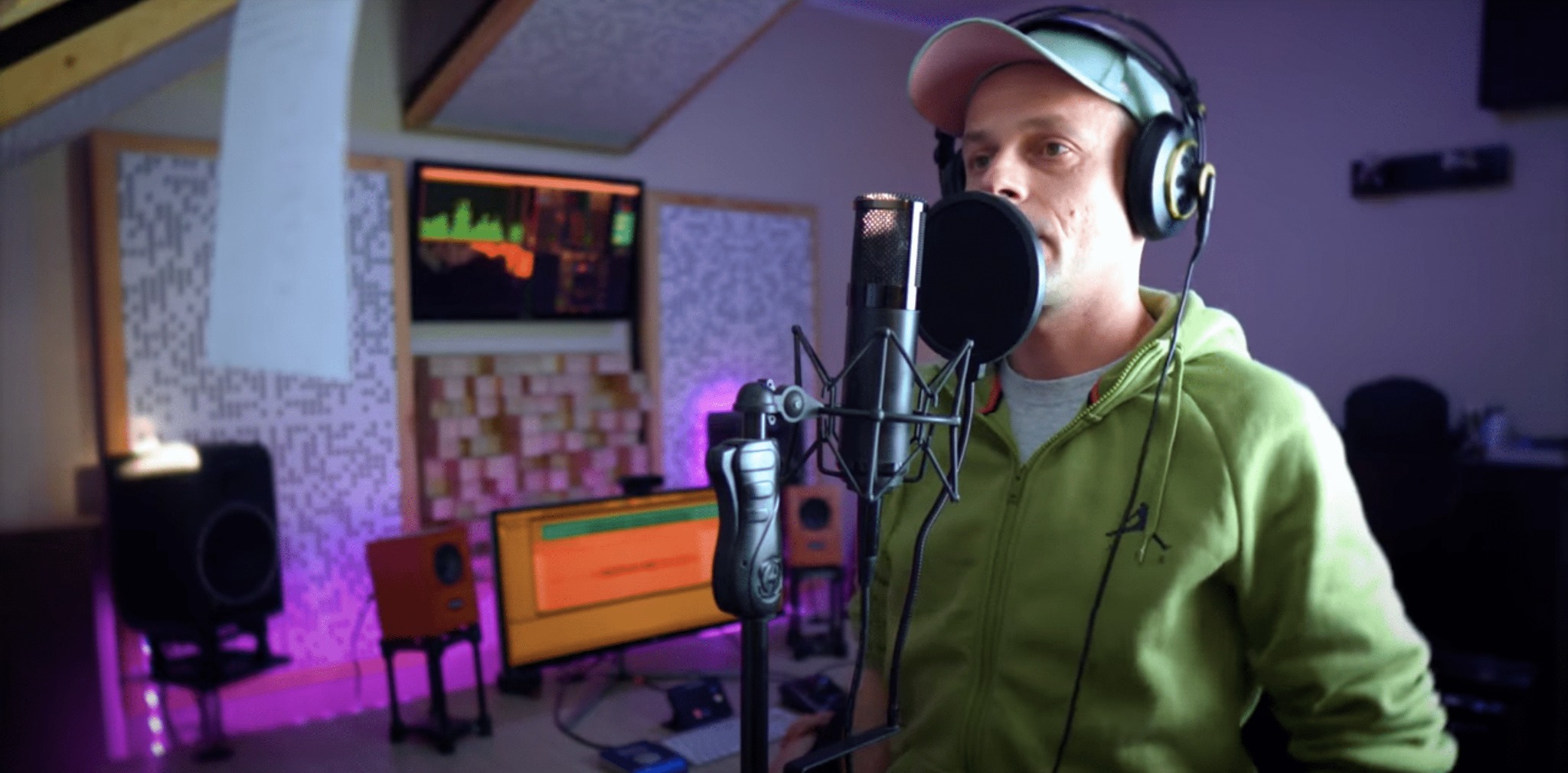 Mężczyzna w słuchawkach i czapce nagrywa wokal w profesjonalnym studiu nagrań, z widocznym mikrofonem pojemnościowym, ekranem komputera z oprogramowaniem DAW, głośnikami studyjnymi i panelami...