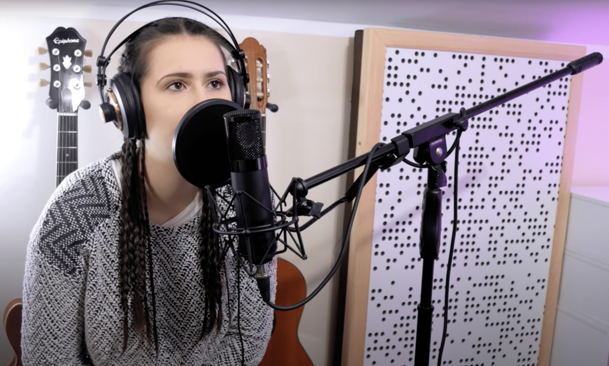 Wokalistka z warkoczami śpiewa do mikrofonu w studiu, w tle gitary i panel akustyczny z otworami.