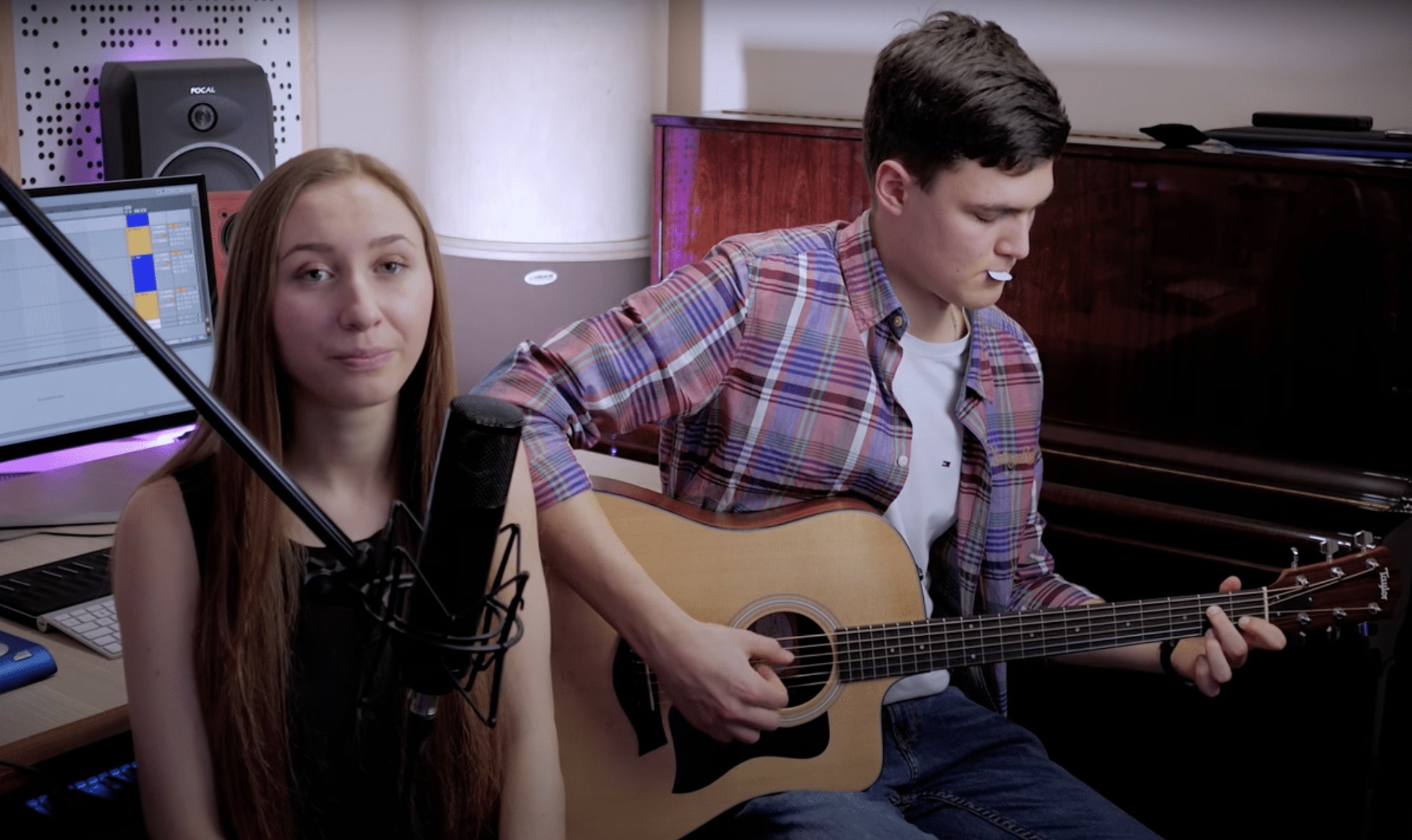 Duet muzyczny w domowym studiu: młoda kobieta śpiewa do mikrofonu, a mężczyzna gra na gitarze akustycznej, mając w ustach kostkę do gitary. W tle monitor komputera z programem do nagrywania i pianino.
