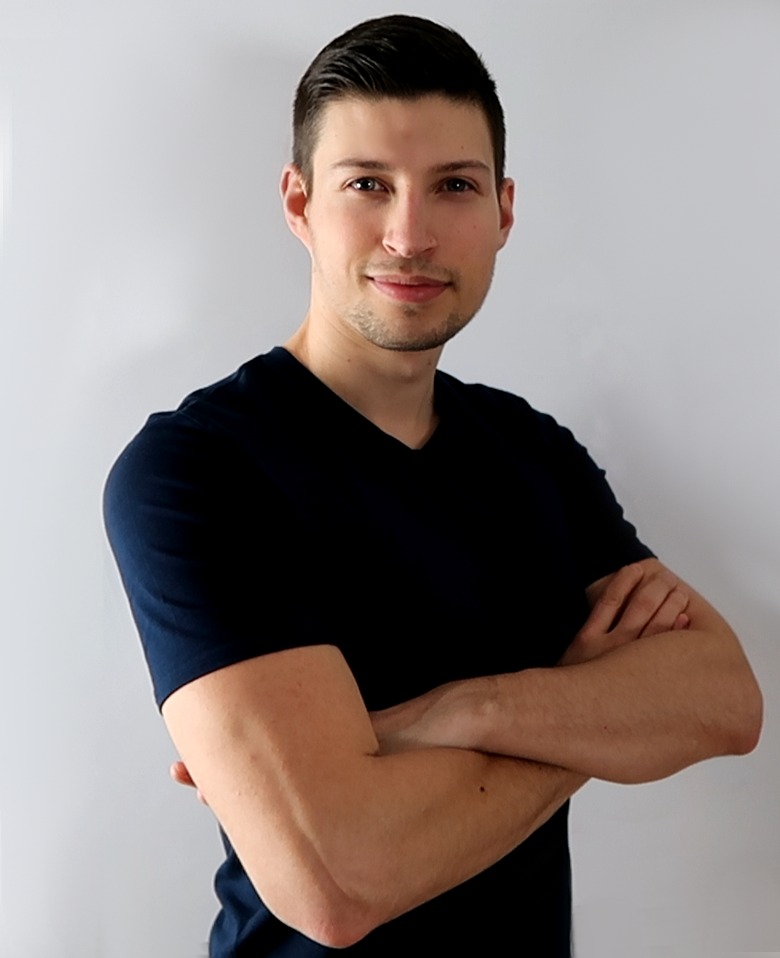 Mężczyzna w ciemnej koszulce z rękawami do łokcia, z założonymi rękami, uśmiecha się, stojąc na tle białej ściany. Widoczny cień na ścianie za nim.