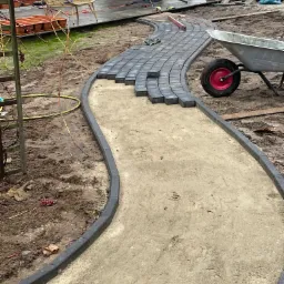 Krzywoliniowy chodnik z szarej kostki brukowej w trakcie układania, z widocznymi obrzeżami, piaskowym podłożem, taczką z czerwoną felgą i poziomnicą na ułożonej części, w tle dom i elementy...