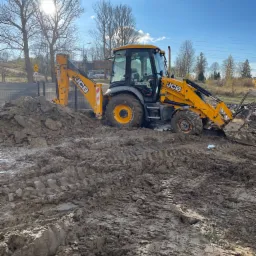 Żółta koparko-ładowarka JCB zanurzona w błotnistym terenie, widoczne ślady opon, w tle drzewa i budynki pod błękitnym niebem z promieniami słońca.