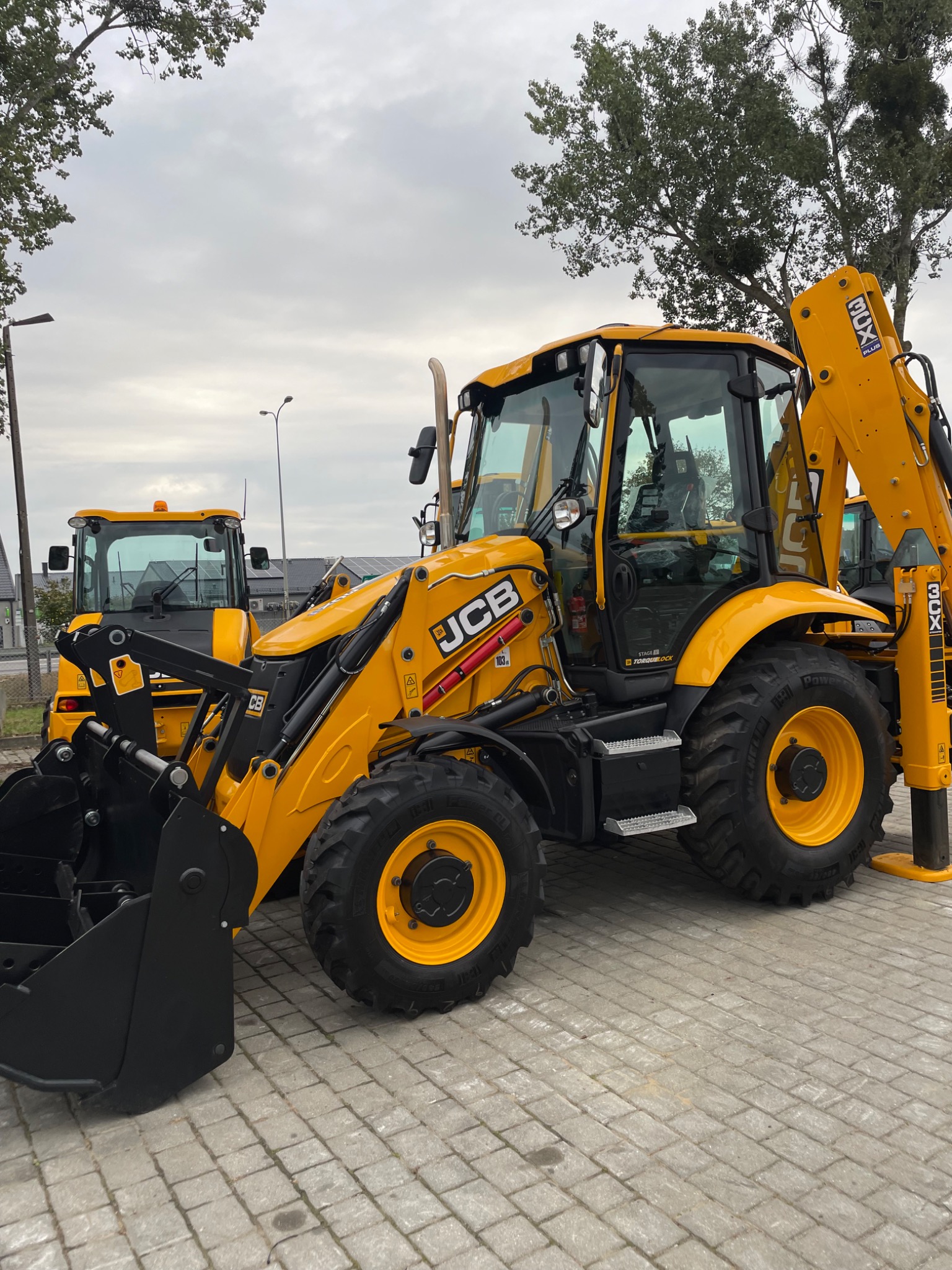 Żółta koparko-ładowarka JCB 3CX na kostce brukowej, z widoczną łyżką i ramieniem koparki, w tle inne maszyny i drzewa pod pochmurnym niebem.