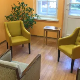 Jasne pomarańczowe wnętrze gabinetu z trzema fotelami (dwa żółte, jeden szary) ustawionymi w kręgu, mały drewniany stolik przy oknie i roślina doniczkowa.