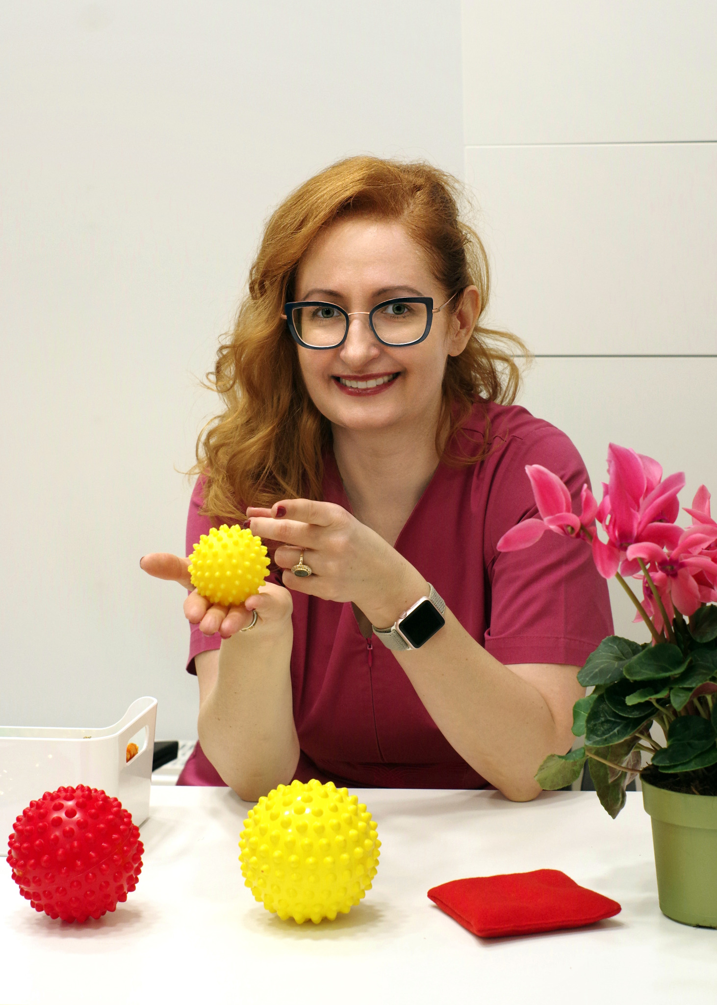 Uśmiechnięta kobieta w okularach i bordowym uniformie prezentuje żółtą piłeczkę sensoryczną na tle białego gabinetu z kwiatem w doniczce i dwoma piłeczkami na stole.