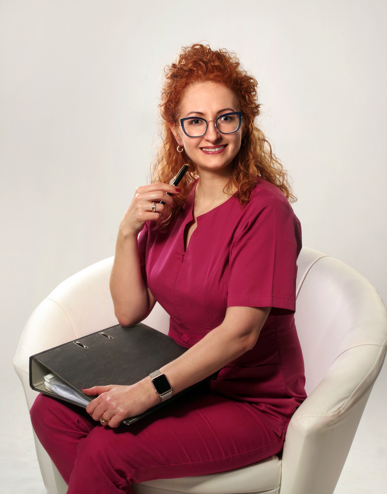 Uśmiechnięta kobieta w okularach i bordowym uniformie siedzi w białym fotelu, trzymając czarny segregator z dokumentami i długopis w dłoni.