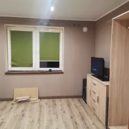 Pokój w trakcie urządzania: widoczne panele podłogowe, okno z zielonymi roletami, szafka RTV z telewizorem, głośnik, częściowo złożona szafa i elementy do montażu mebli pod oknem.