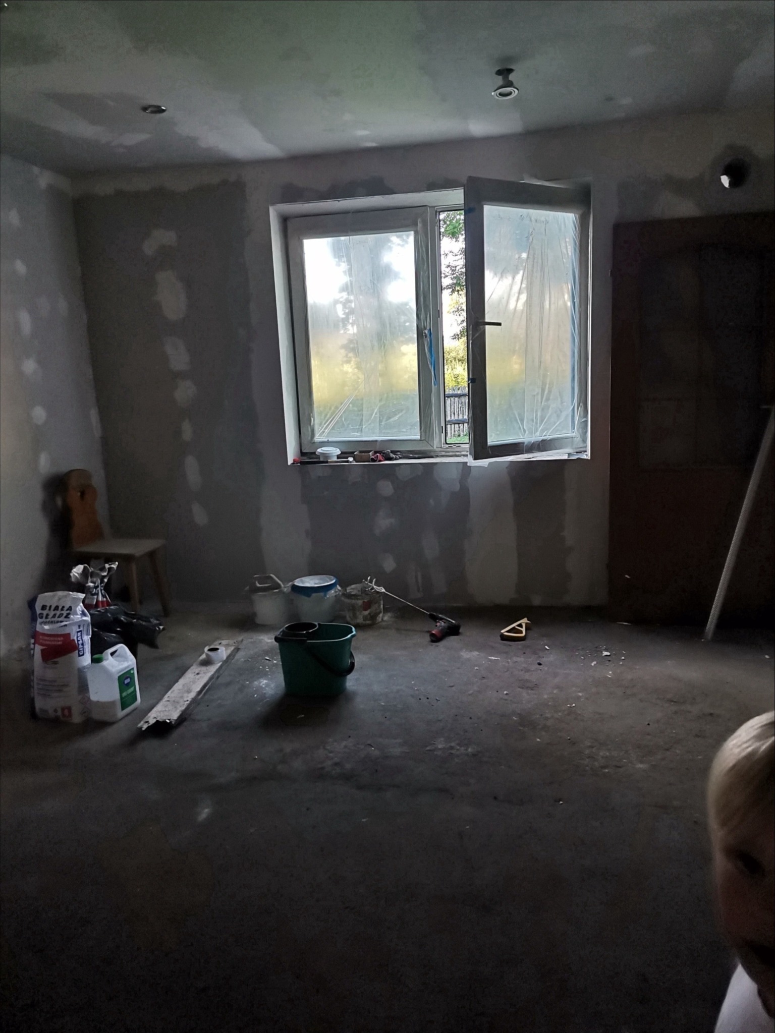 Pokój w trakcie prac wykończeniowych: ściany z nałożoną gładzią, okno zabezpieczone folią, widoczne narzędzia i materiały budowlane, w tle krzesło i drzwi.