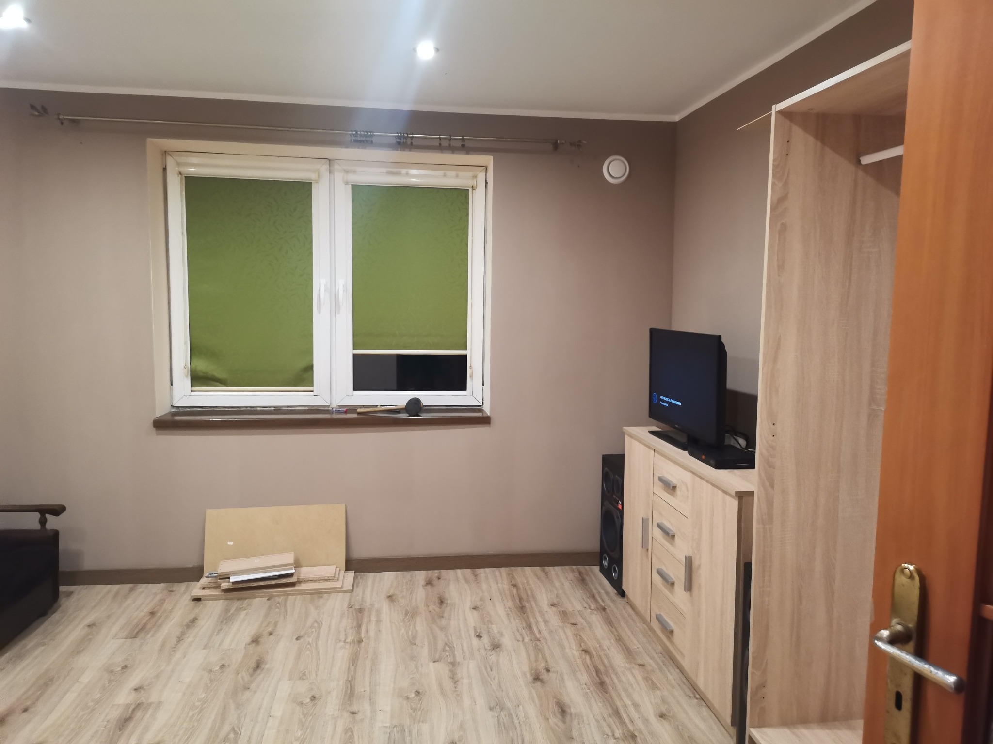 Pokój w trakcie urządzania: widoczne panele podłogowe, okno z zielonymi roletami, szafka RTV z telewizorem, głośnik, częściowo złożona szafa i elementy do montażu mebli pod oknem.