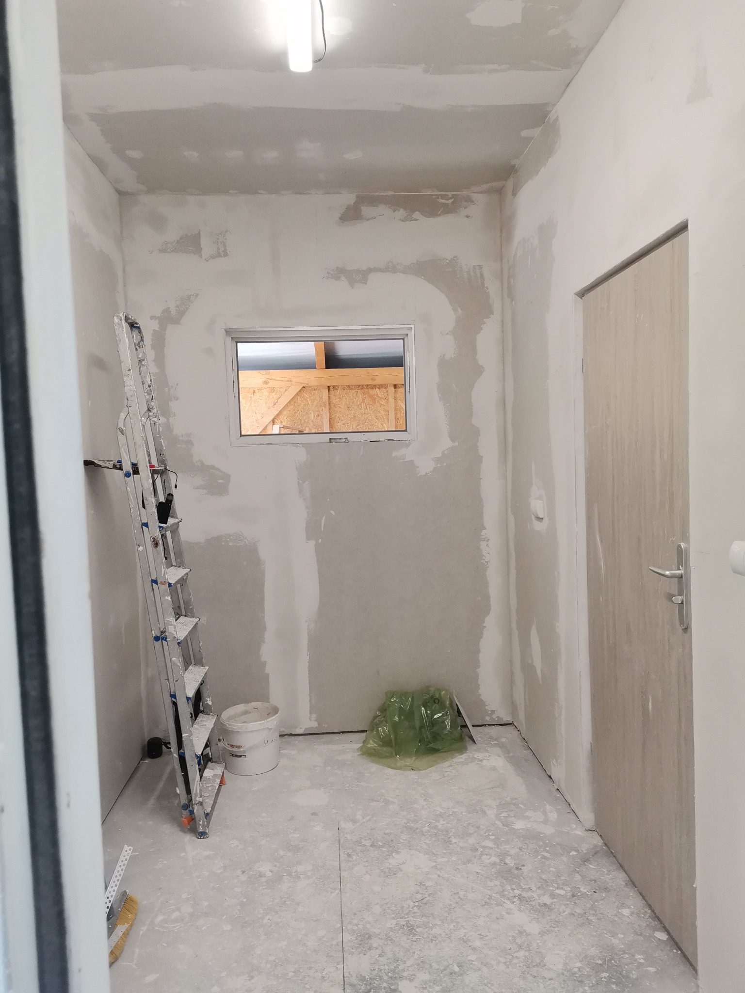 Wnętrze w trakcie prac wykończeniowych: ściany z nałożoną, częściowo wyschniętą gładzią, aluminiowa drabina oparta o ścianę, okno z widoczną konstrukcją drewnianą na zewnątrz, drzwi wewnętrzne...