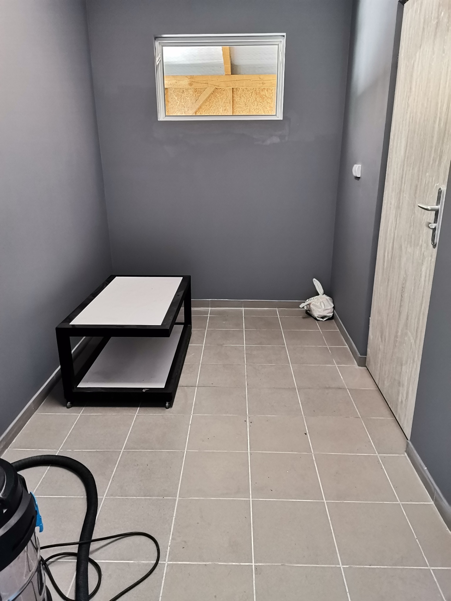 Szare pomieszczenie po remoncie z nowymi płytkami podłogowymi, widoczne ślady po pracy, okno z widokiem na konstrukcję dachu, stolik na kółkach i odkurzacz przemysłowy.