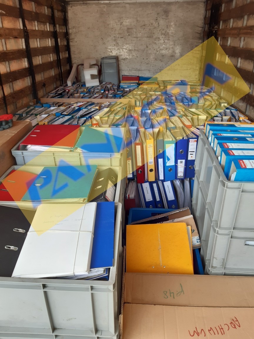 Wnętrze furgonetki wypełnionej po brzegi szarymi plastikowymi pojemnikami i kartonami, w których znajdują się segregatory z dokumentami o różnych kolorach, przygotowane do transportu.