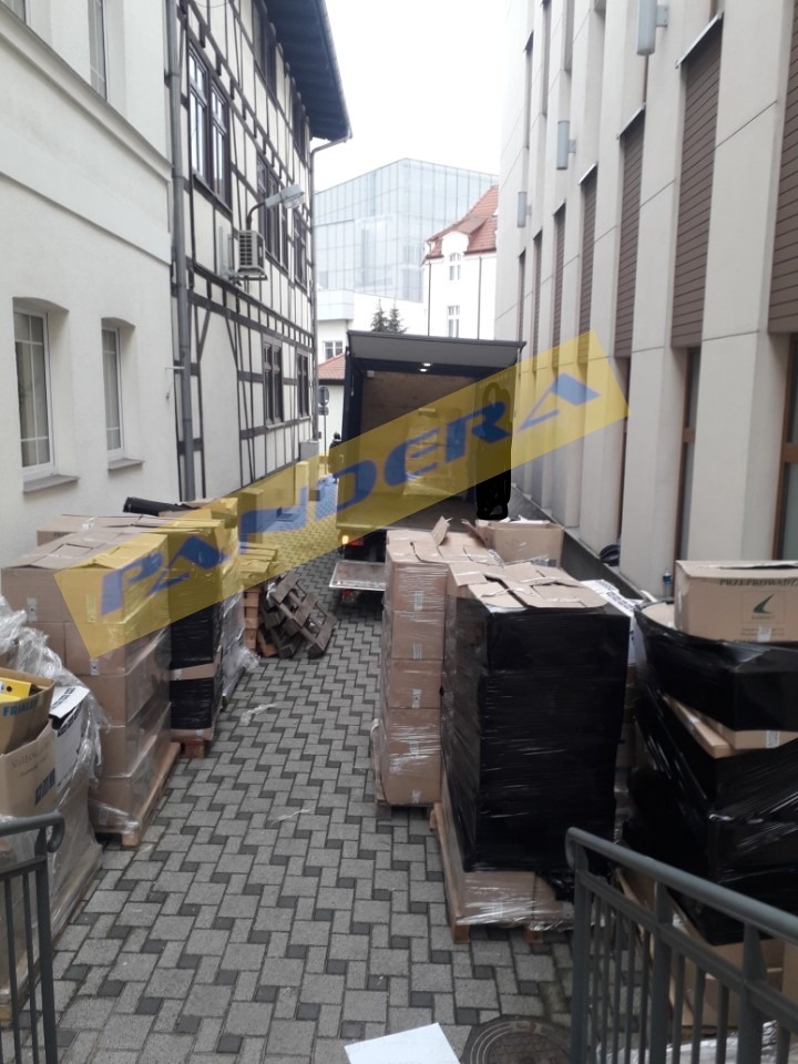 Załadunek kartonów i palet do samochodu ciężarowego zaparkowanego pomiędzy budynkami o różnej architekturze, widok z góry schodów.