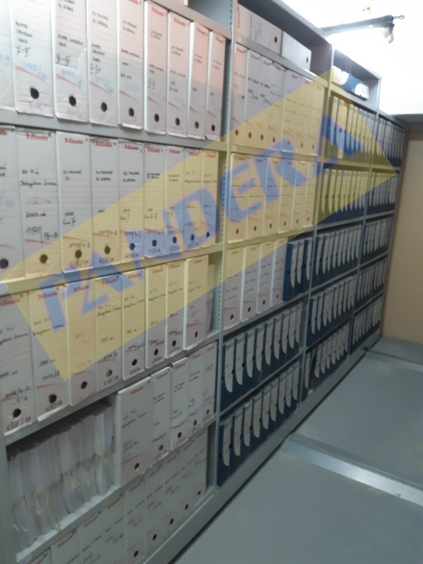 Metalowe regały archiwalne wypełnione segregatorami z dokumentami, widoczne etykiety z datami i numerami akt.