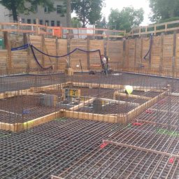 M.K. CONSTRUCTION Michał Kudełka - Siatka zbrojeniowa fundamentu na placu budowy, widoczne drewniane szalunki i pracownicy w tle.