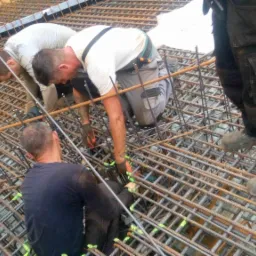 Trzech zbrojarzy w trakcie pracy nad siatką zbrojeniową fundamentu, widoczne elementy szalunku i narzędzia.