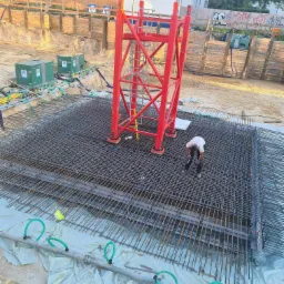 Przygotowanie zbrojenia płyty fundamentowej pod konstrukcję żelbetową; widoczny pracownik w trakcie pracy, czerwona konstrukcja wsporcza, oraz system rur do ogrzewania podłogowego.