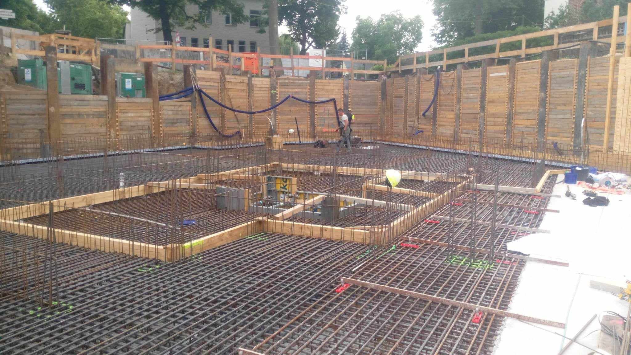 Siatka zbrojeniowa fundamentu na placu budowy, widoczne drewniane szalunki i pracownicy w tle.