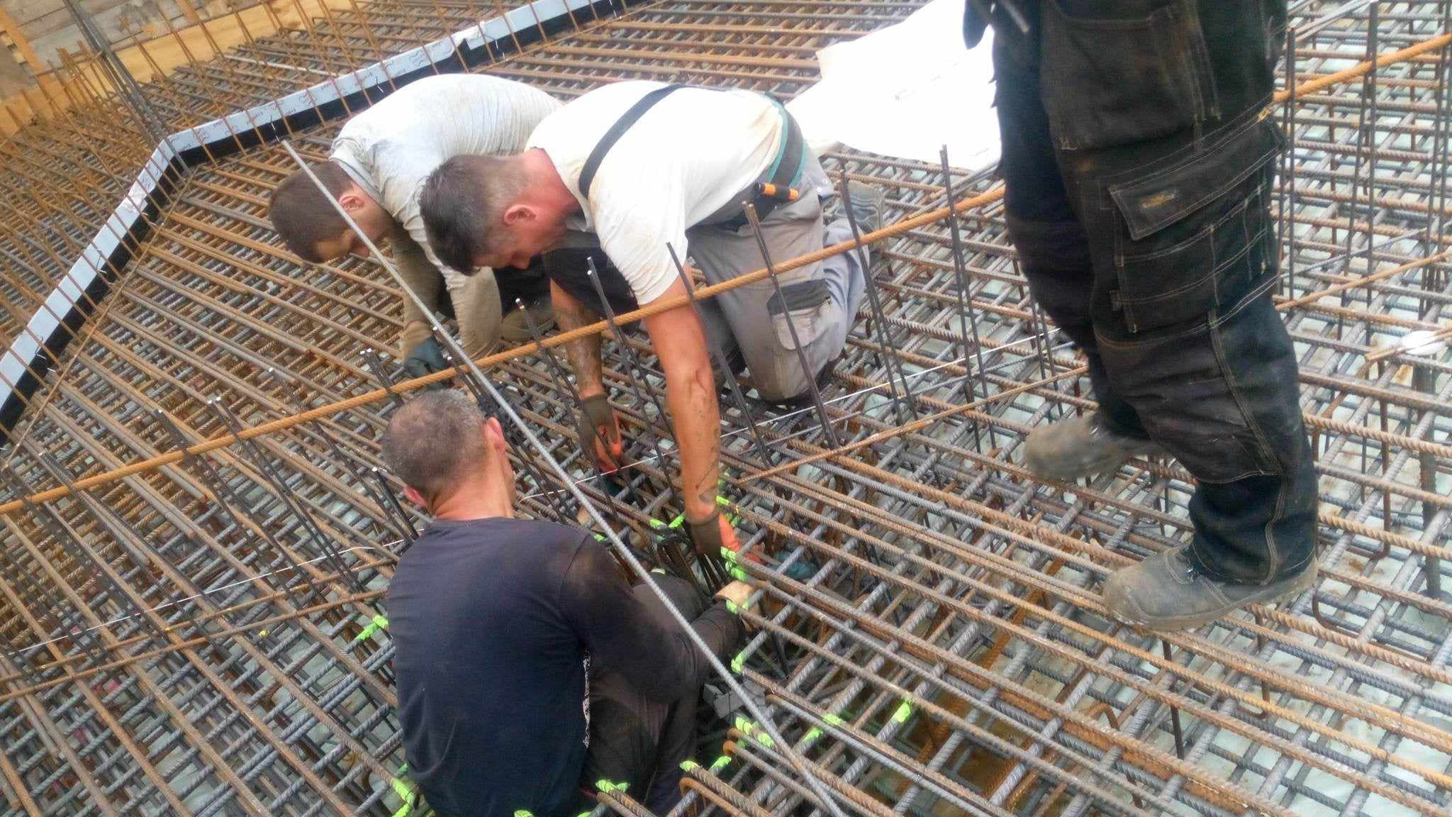 Trzech zbrojarzy w trakcie pracy nad siatką zbrojeniową fundamentu, widoczne elementy szalunku i narzędzia.