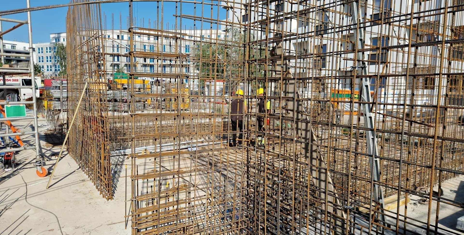 Siatka zbrojeniowa na placu budowy, widoczni pracownicy w żółtych kaskach, w tle budynki mieszkalne.