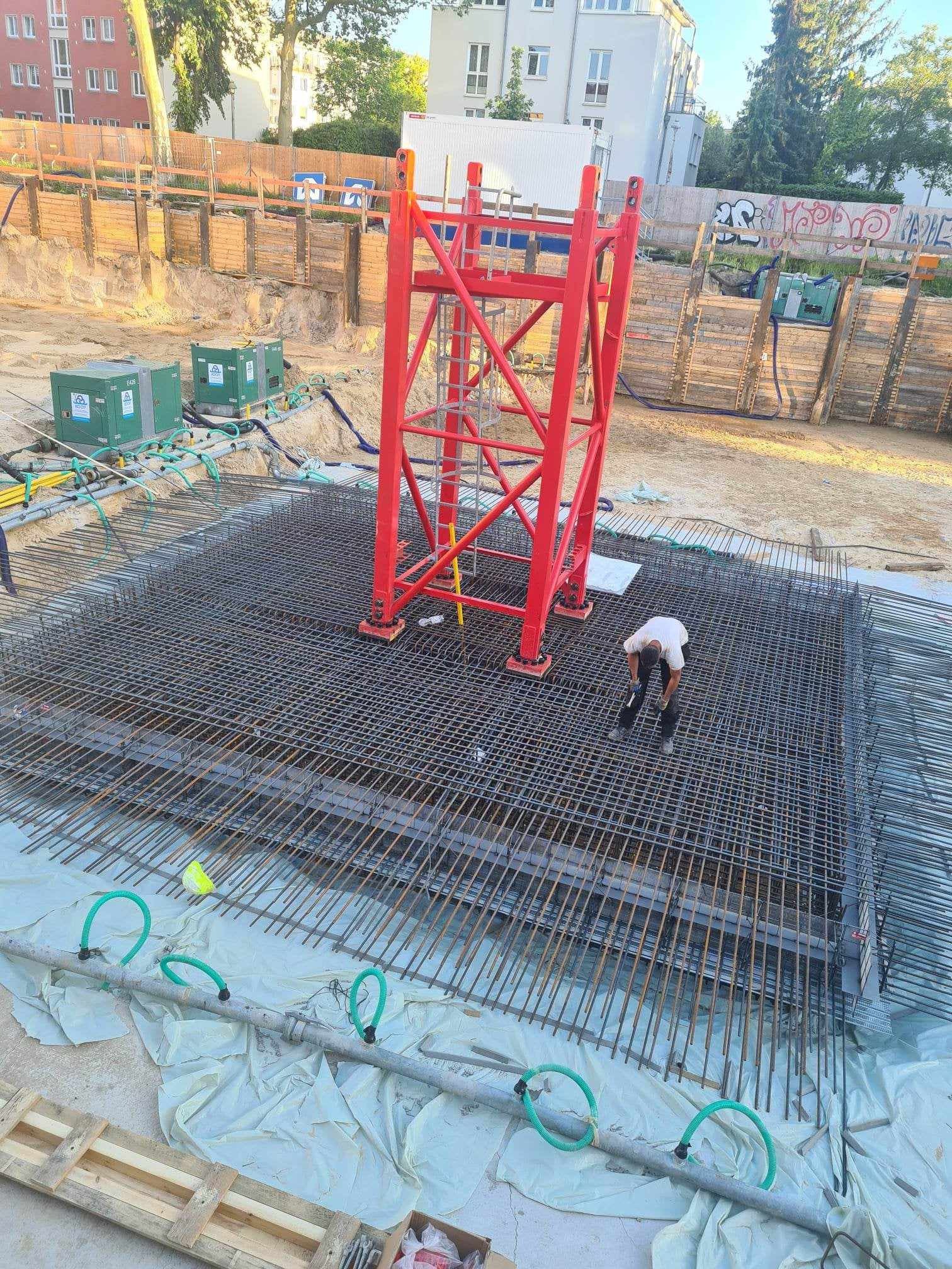 Przygotowanie zbrojenia płyty fundamentowej pod konstrukcję żelbetową; widoczny pracownik w trakcie pracy, czerwona konstrukcja wsporcza, oraz system rur do ogrzewania podłogowego.