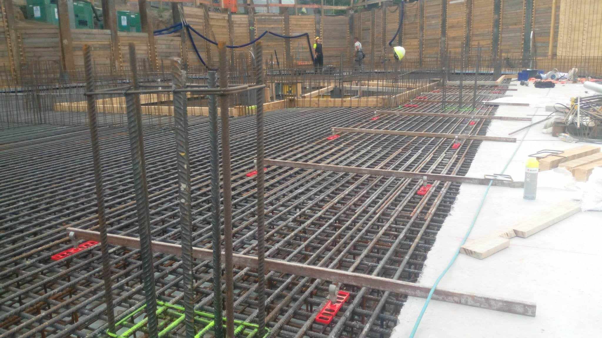 Siatka zbrojeniowa fundamentu z widocznymi dystansami z tworzywa sztucznego, drewniane szalunki i pracownicy w tle.