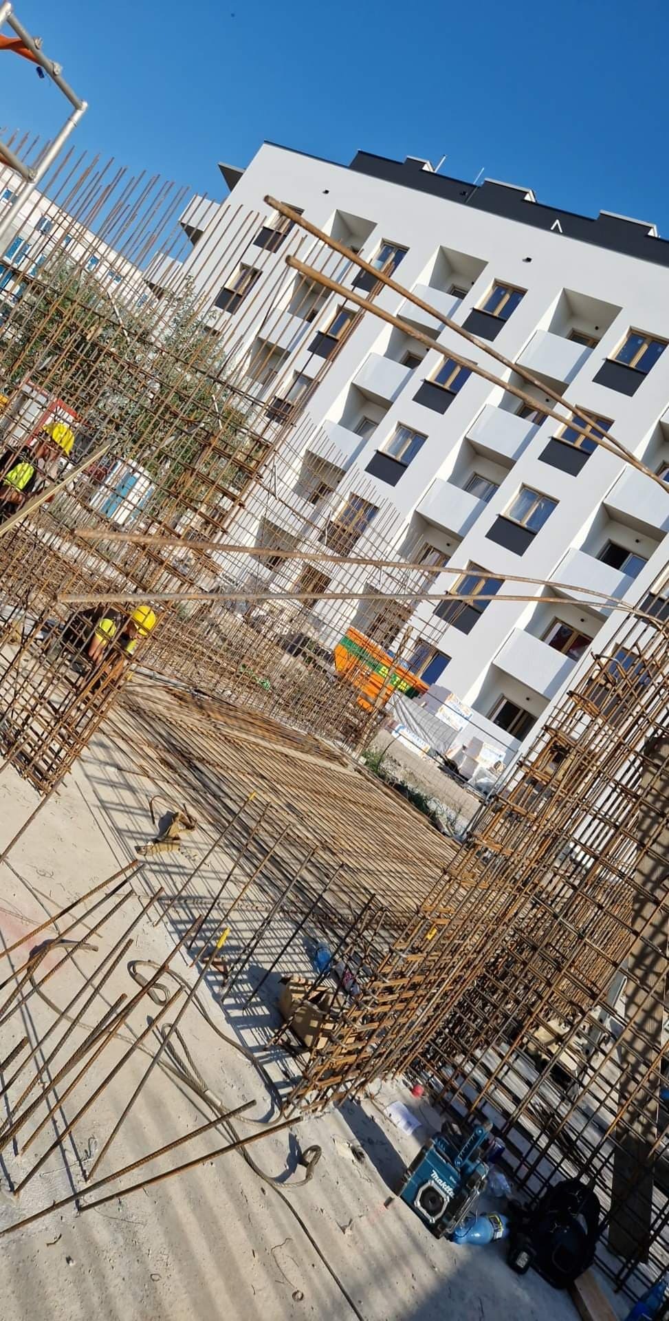 Konstrukcja zbrojeniowa na placu budowy z widocznymi pracownikami w żółtych kaskach, w tle biały budynek mieszkalny z balkonami.