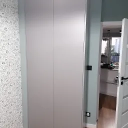 Szara szafa wnękowa z dwiema parami drzwi, na tle ściany z tapetą w czarno-białe rysunki psów, widok na otwarte drzwi do pokoju.