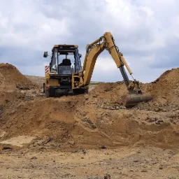 Żółta koparko-ładowarka na nierównym, piaszczystym terenie z wyraźnymi śladami opon, z hałdami ziemi w tle i stogiem siana przykrytym plandeką po lewej stronie, pod pochmurnym niebem.