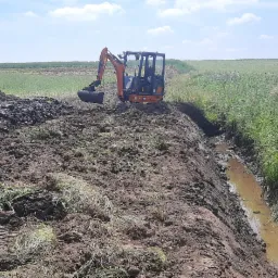 Pomarańczowa mini koparka Hitachi na skraju wykopu wypełnionego wodą, widok na pole i drzewa w tle.