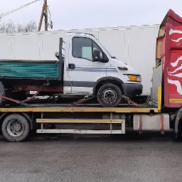 Laweta z czerwono-białym wzorem transportuje białą wywrotkę z zieloną skrzynią ładunkową, zabezpieczoną pasami transportowymi.