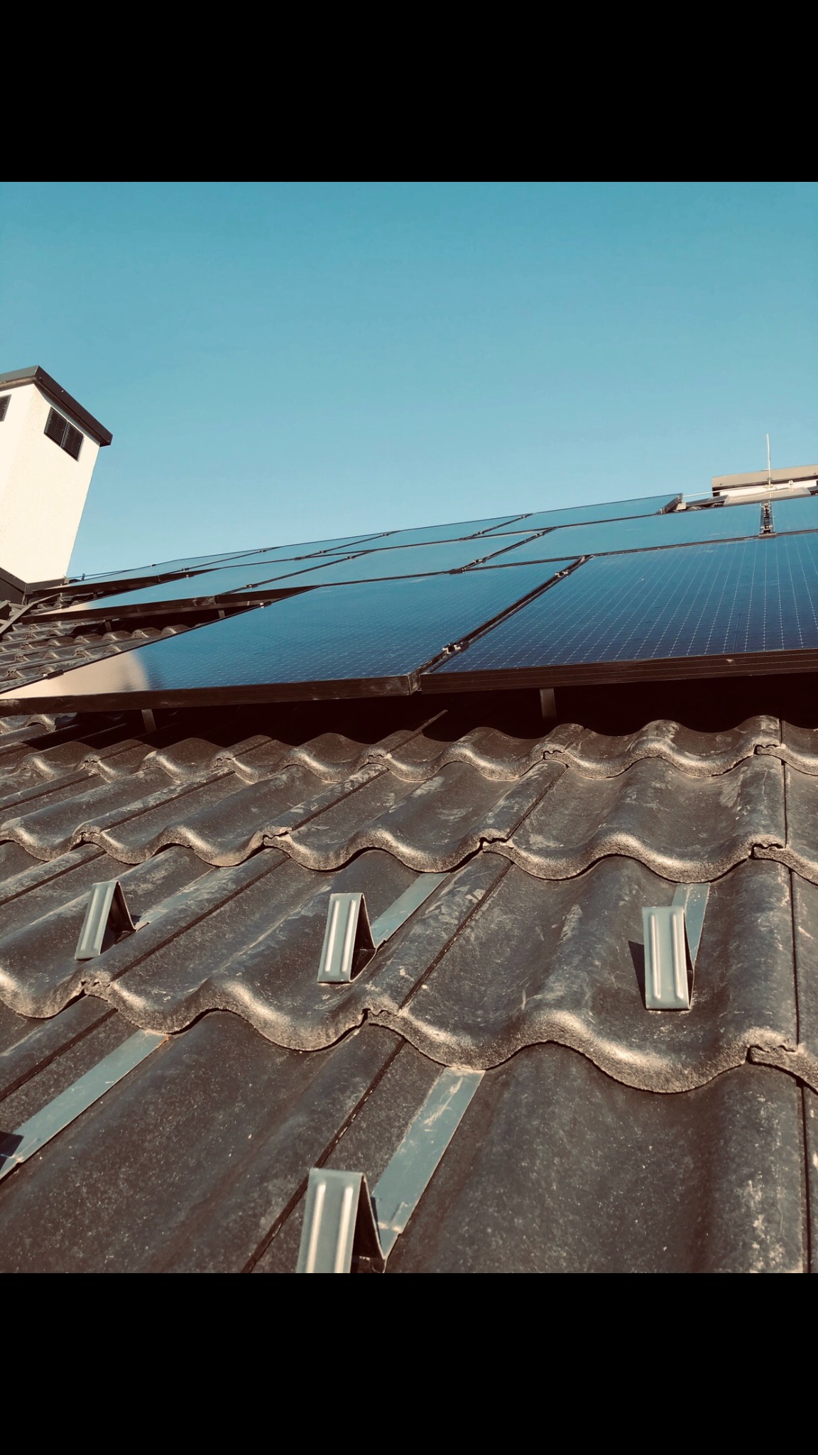 Panele fotowoltaiczne zamontowane na ciemnej, ceramicznej dachówce z widocznymi metalowymi uchwytami montażowymi, błękitne niebo w tle.