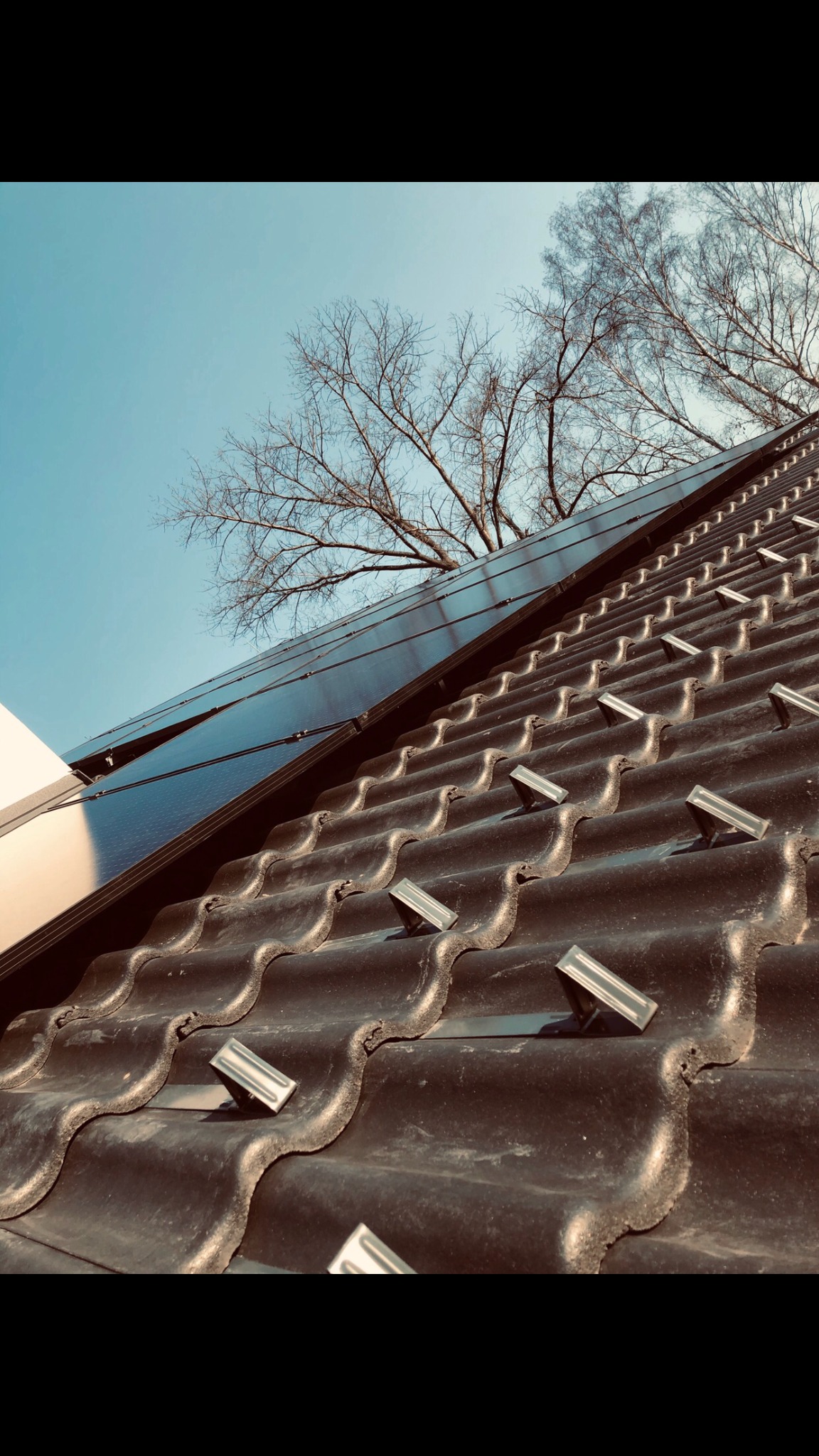 Ciemne panele słoneczne zamontowane na brązowym, ceramicznym dachu z widocznymi mocowaniami, na tle błękitnego nieba i gałęzi drzew bez liści.