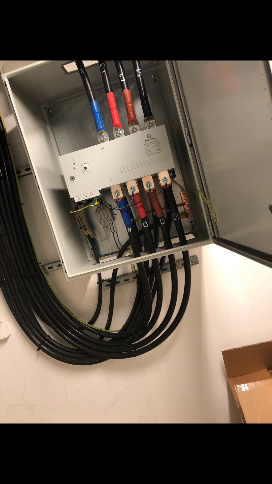 Otwarta skrzynka elektryczna z podłączonymi grubymi kablami oznaczonymi kolorami i symbolami N, L1, L2, L3, widoczne elementy montażowe na szynie DIN.