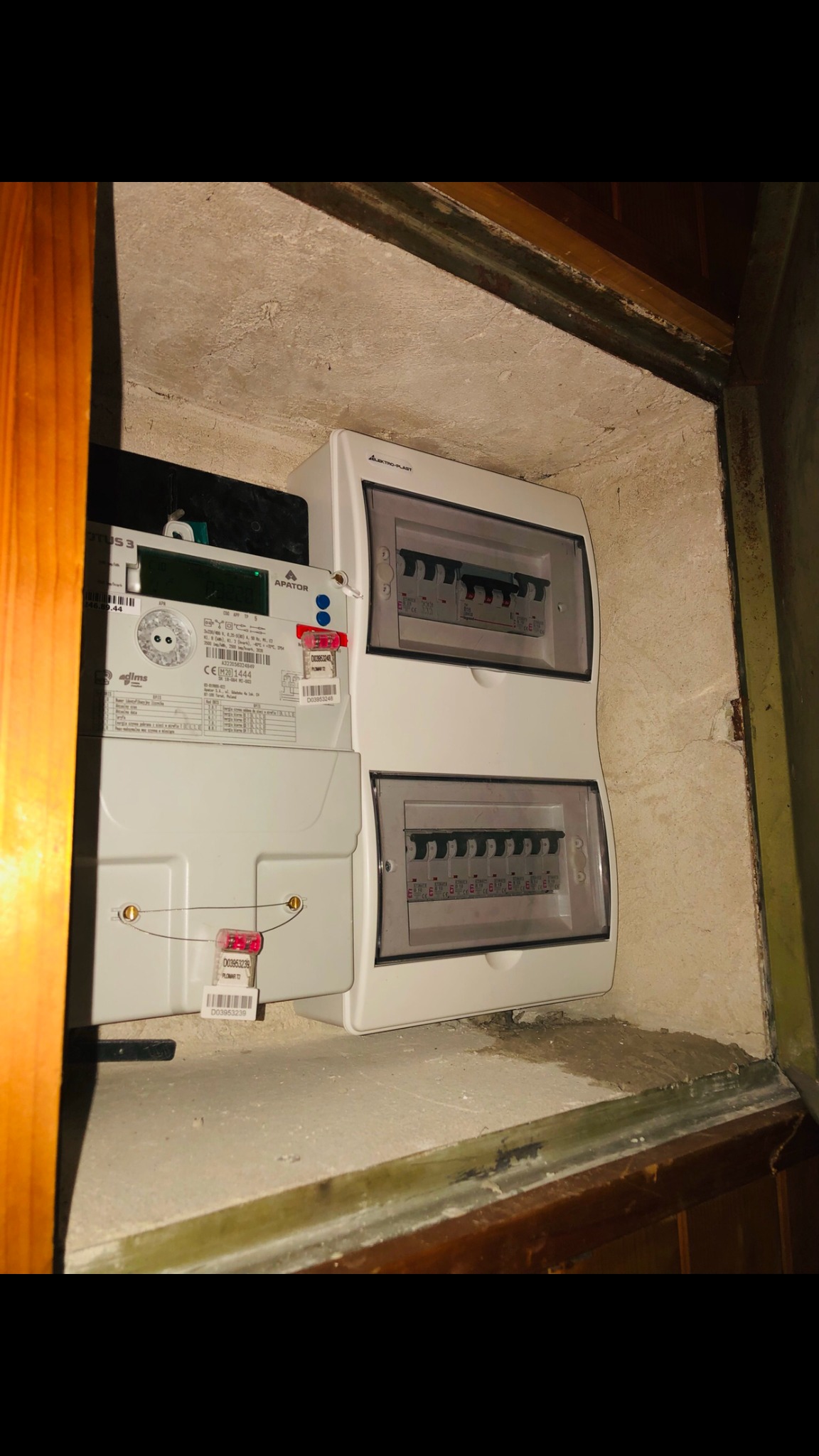 Nowy, biały licznik energii elektrycznej i dwie rozdzielnice w drewnianej wnęce, zabezpieczone plombami.