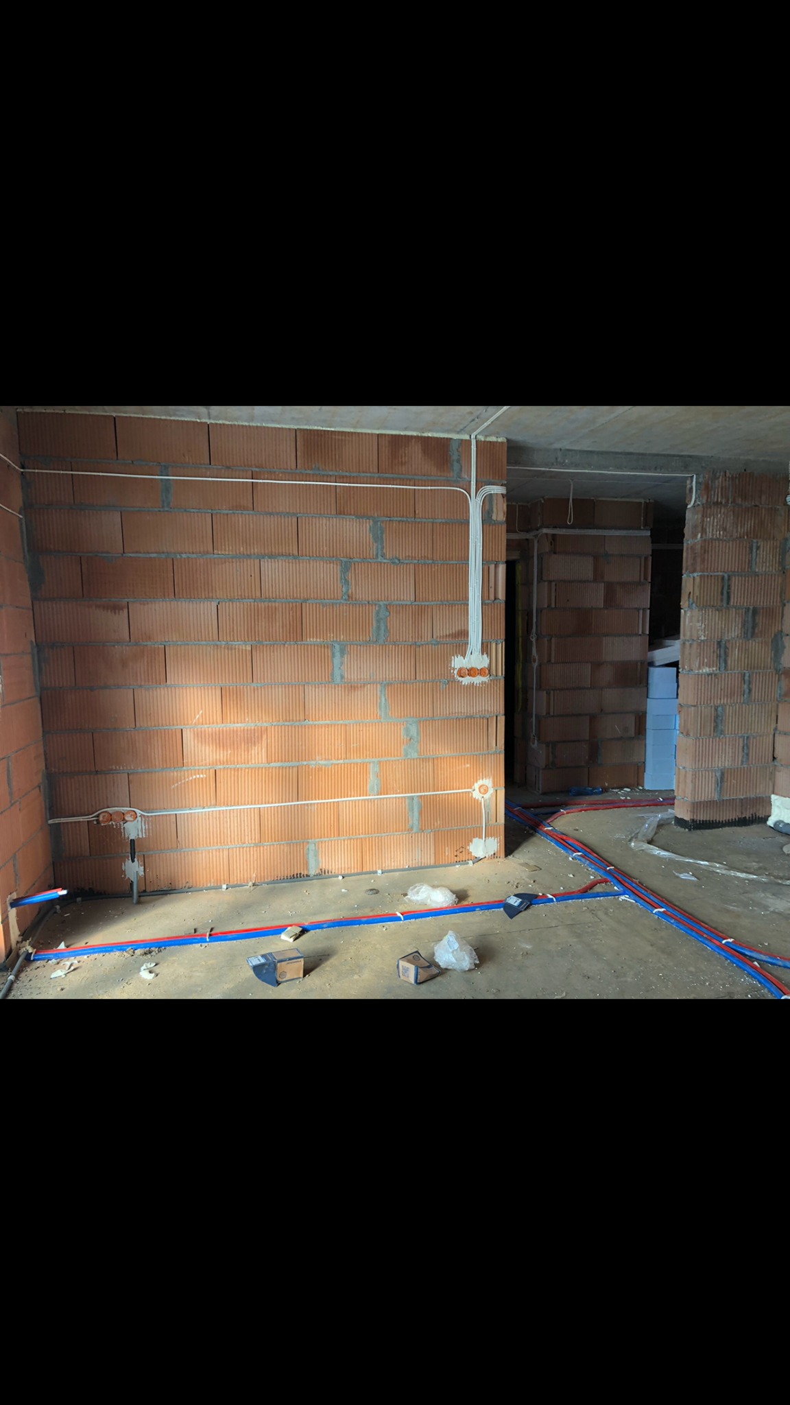 Wnętrze w trakcie instalacji elektrycznej i hydraulicznej: ceglane ściany, białe kable elektryczne prowadzone po ścianach, niebiesko-czerwone rury na podłodze, puszki instalacyjne i gruz.