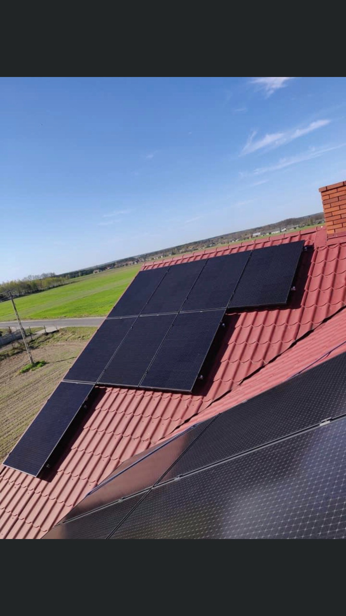 Panele słoneczne zamontowane na czerwonym dachu domu jednorodzinnego, widok z góry na tle zielonego pola i błękitnego nieba.