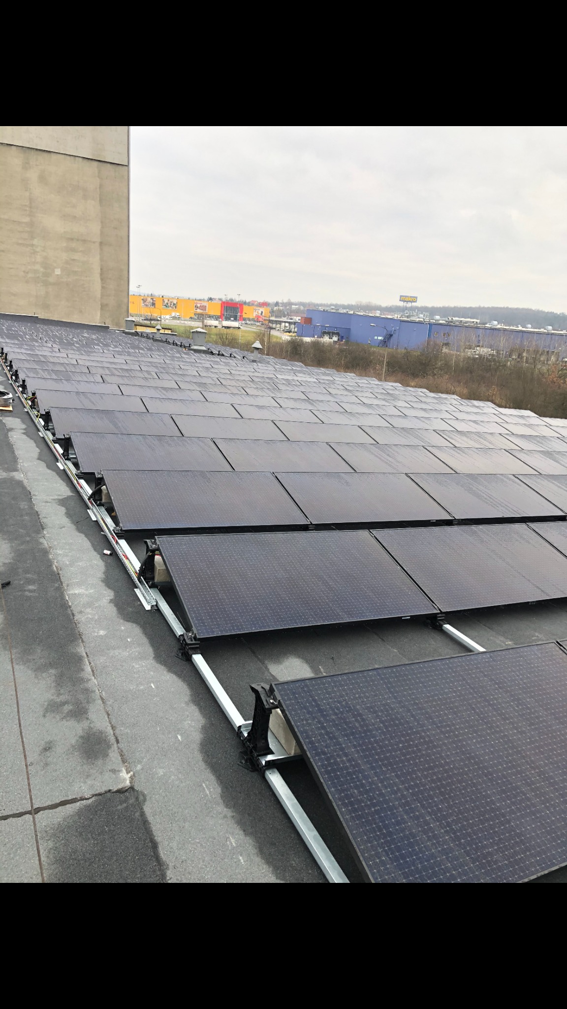 Rząd paneli słonecznych na płaskim dachu z widocznymi elementami montażowymi, w tle zabudowania handlowe i pochmurne niebo.