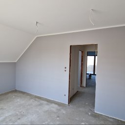 KRIS MAR - Pomalowane na szaro wnętrze poddasza ze skośnym sufitem, widoczne wystające kable elektryczne i surowa podłoga, z otwartym przejściem do kolejnego pomieszczenia.