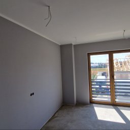 KRIS MAR - Pokój po malowaniu na szaro, widoczne wystające kable ze ściany i sufitu, surowa podłoga, okno balkonowe z widokiem na budowę domu.