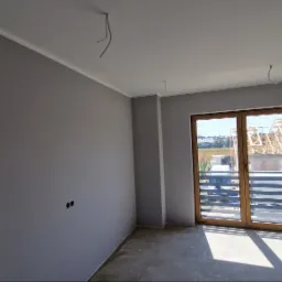 Pokój po malowaniu na szaro, widoczne wystające kable ze ściany i sufitu, surowa podłoga, okno balkonowe z widokiem na budowę domu.